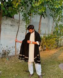 Man in white traditional outfit with black scarf standing outdoors near greenery.